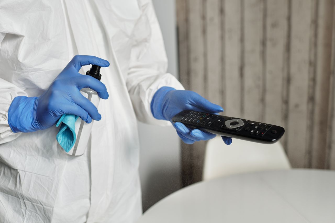 Hands wearing gloves disinfecting a remote control indoors. Pandemic hygiene concept.