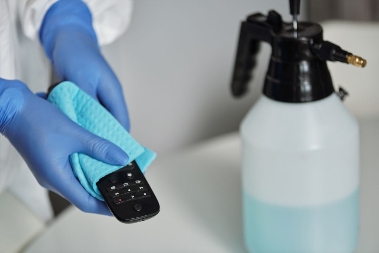 Close-up view of gloved hands sanitizing a remote control with a cloth and spray bottle.